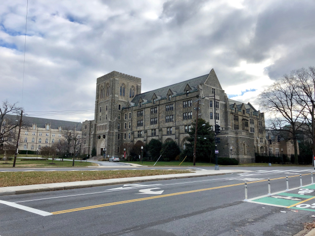 Großes Gebäude mit vielen Fenstern, als Universität von Pennsylvania beschriftet, umgeben von Bäumen, Straßenlaternen, Verkehrszeichen und Fahrzeugen unter einem bewölkten Himmel.