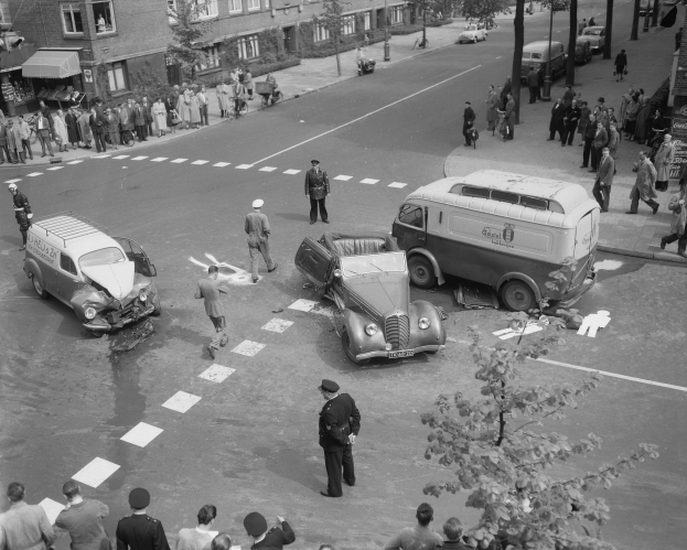 Eine schwarz-weiße Szenerie eines Autounfalls an einer Straßenecke, mit mehreren Fahrzeugen, darunter ein Lieferwagen, einer Gruppe von Menschen drumherum und im Hintergrund Gebäude, Bäume, Masten und Texttafeln.