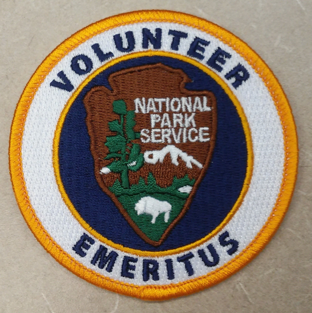 Ein rundes Patch mit einem blauen Rand und einem weißen Hintergrund, das in der Mitte schwarze Schrift 'Volunteer National Park Service Emeritus' in einem gelben Kreis zeigt.