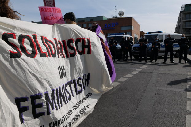 Gruppe von Menschen, die mit einem "Solidarität und Feminismus"-Schild die Straße entlangmarschieren, mit geparkten Fahrzeugen, Gebäuden, einer Schüsselantenne und einem klaren blauen Himmel im Hintergrund.