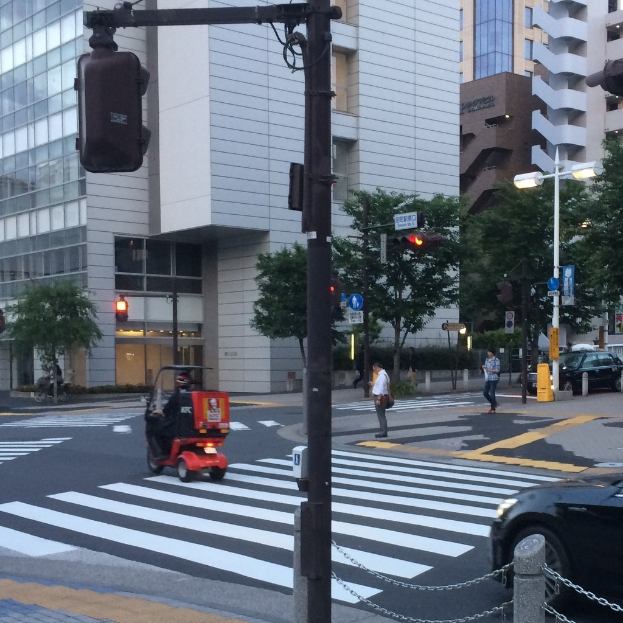 Ein E-Scooter auf einer Straße mit Menschen im Hintergrund, eine Ampel im Vordergrund und Gebäude mit Bäumen im Hintergrund.