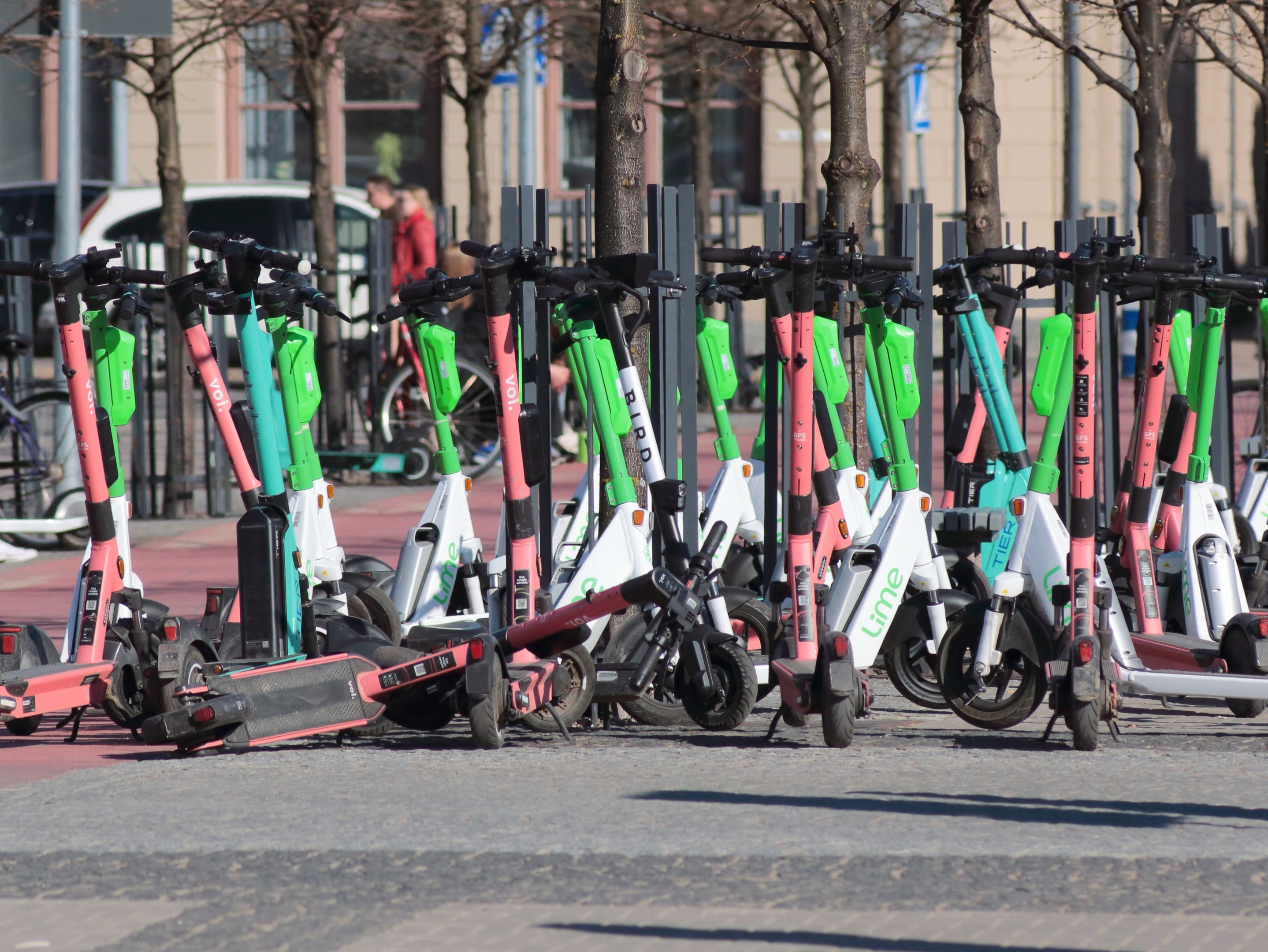 Eine Reihe von Elektro-Rollern, die an einer Straße geparkt sind, mit einer Person im Vordergrund, Bäumen, Masten, Schildern, Fahrzeugen und Gebäuden im Hintergrund.