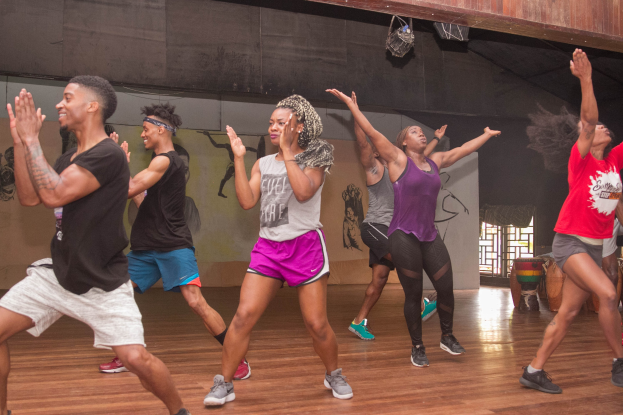 Eine Gruppe von Menschen, die in einem Studio mit einer bemalten Wand, einem Fenster und Deckenbeleuchtung tanzen, wahrscheinlich während eines Zumba-Kurses.