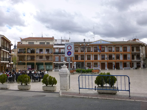 Ein belebter Stadtplatz mit sitzenden und stehenden Menschen, Topfpflanzen, Metallabsperrungen, einem Schild an einem Pfahl, Straßenlaternen mit Flaggen, umliegenden Gebäuden mit Fenstern und einem bewölkten Himmel.