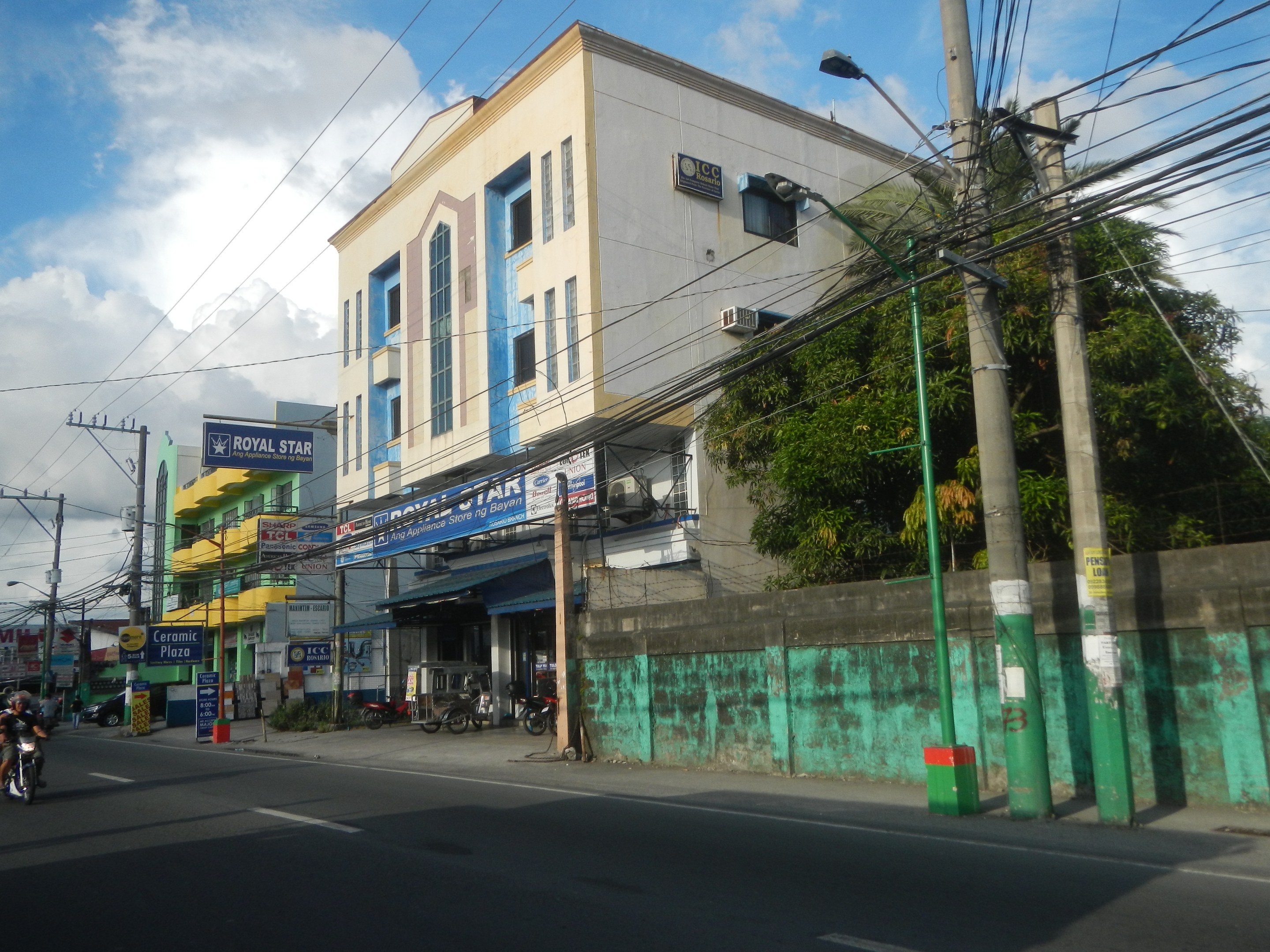 Stadtstraßenszene mit Gebäuden, Fahrzeugen, Strommasten, Kabeln, Straßenlaternen, Schildern, Bäumen und bewölktem Himmel, mit einem Gebäude im Vordergrund, das dicht mit Kabeln bedeckt ist.