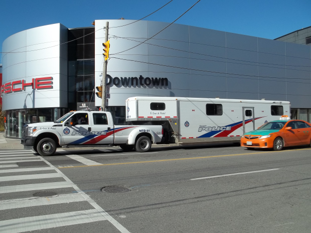 Ein Polizeiwagen steht vor einem Gebäude mit Text darauf, mit einer Verkehrsampel mit Stange und Drähten im Vordergrund und Bäumen und einem klaren blauen Himmel im Hintergrund.