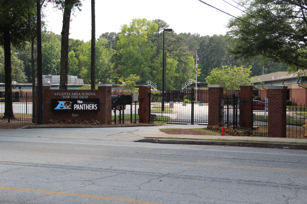 Der Eingang einer Schule mit einem Schild an der Seite, umgeben von Bäumen, Straßenlaternen, Gittern, Namensschildern, Fahrzeugen auf der Straße, Absperrgittern, Pflanzen, Gras, Gebäuden und einem klaren blauen Himmel.