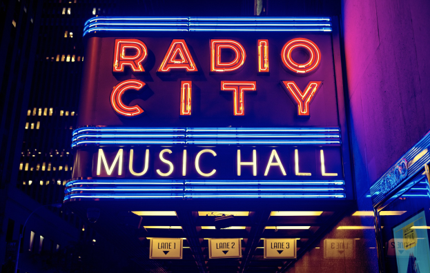 Hell erleuchtetes Radio City Music Hall in New York City mit hohen Gebäuden im Hintergrund und einer 'Radio City'-Leuchtschrift auf der rechten Seite.