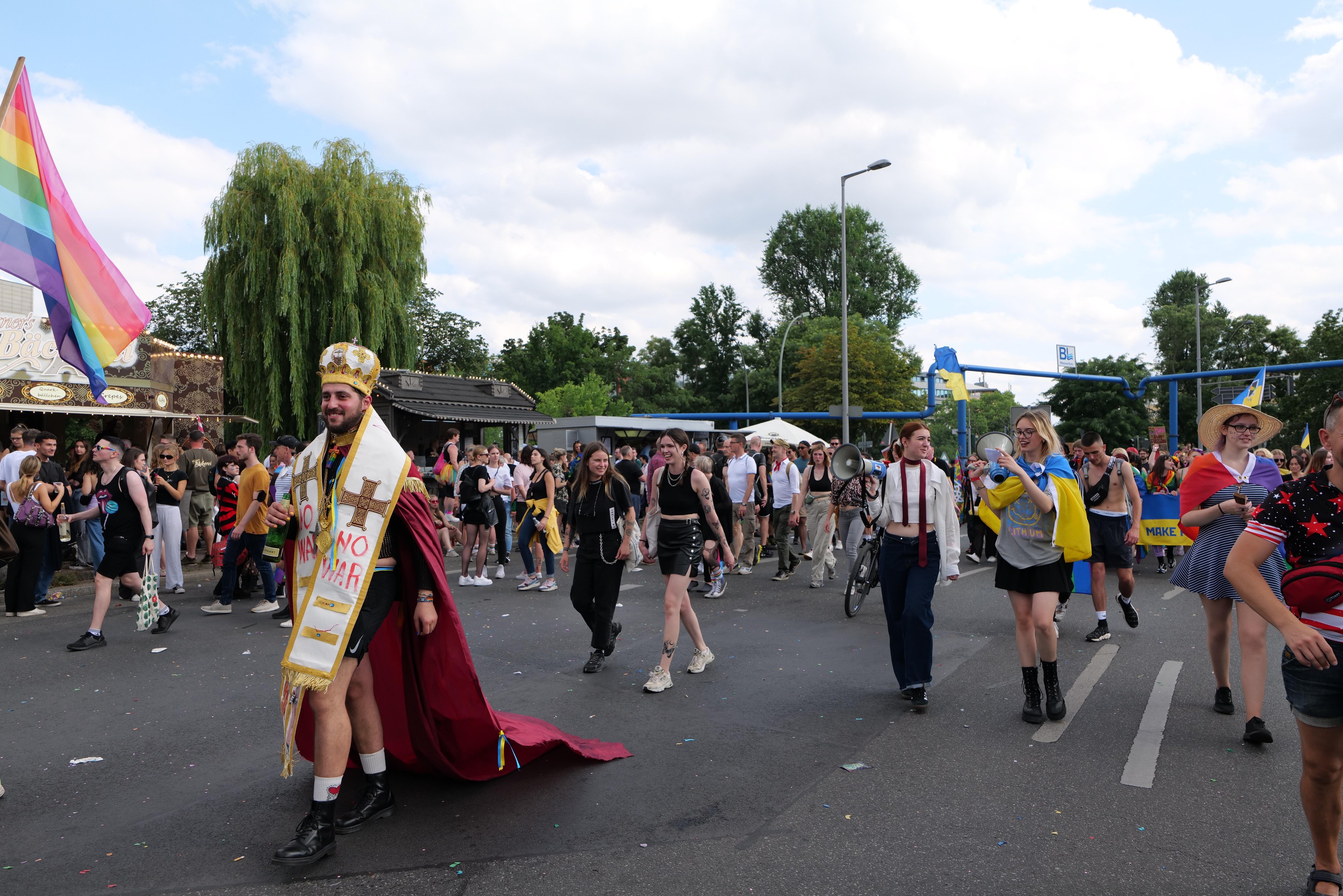Eine Gruppe von Menschen bei der 2018er Gay Pride Parade mit einer Regenbogenflagge und Musikinstrumenten, einige tragen Mützen, vor dem Hintergrund von Laternenpfählen, Bäumen, Schuppen und einem bewölkten Himmel.