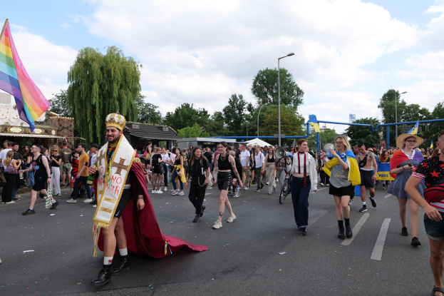 Eine Gruppe von Menschen bei der 2018er Gay Pride Parade mit einer Regenbogenflagge und Musikinstrumenten, einige tragen Mützen, vor dem Hintergrund von Laternenpfählen, Bäumen, Schuppen und einem bewölkten Himmel.
