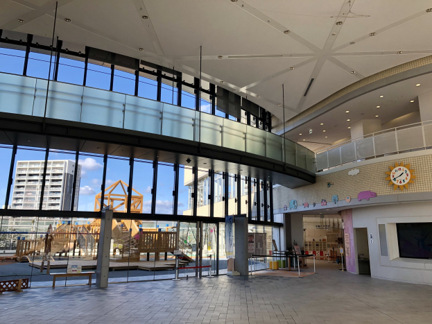 Innenansicht des Haupt входs zu einem neuen Kinderspielbereich mit großen Fenstern, elektrischen Lampen, einer Wanduhr, einem Fernseher, einem Anzeigebildschirm, hölzernen Gegenständen, einem hölzernen Zaun, umgeben von Gebäuden, Bäumen und einem bewölkten Himmel.