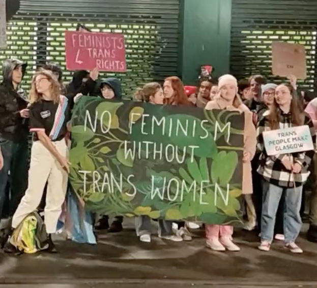 Eine Gruppe von Menschen hält ein Transparent mit der Aufschrift "Kein Feminismus ohne Transfrauen" und verschiedene Schilder in einer öffentlichen Fläche mit einer Wand und einem Rollladen im Hintergrund.