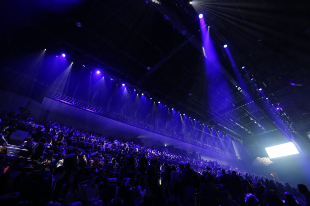 Eine große Menschenmenge sitzt vor einer Bühne mit Deckenleuchten und einem Bildschirm auf der rechten Seite, was auf eine Konzertlocation hindeutet.