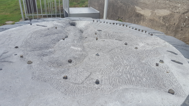 Großer Betonbau mit kreisförmiger Gestaltung oben drauf, umgeben von einem Metallzaun, einem Pfahl, einer Wand und Gras.