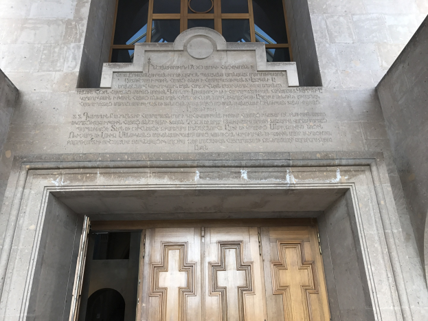 Große hölzerne Tür vor einem steinernen Gebäude mit einem Glasfenster oben, das wahrscheinlich Holocaust-Opfern gewidmet ist.