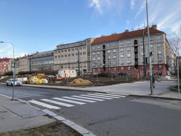 Baustelle für einen neuen Wohnblock im Vordergrund einer städtischen Straße mit parkenden Autos, Laternen, Bäumen, Gebäuden mit Fenstern und einem bewölkten Himmel.