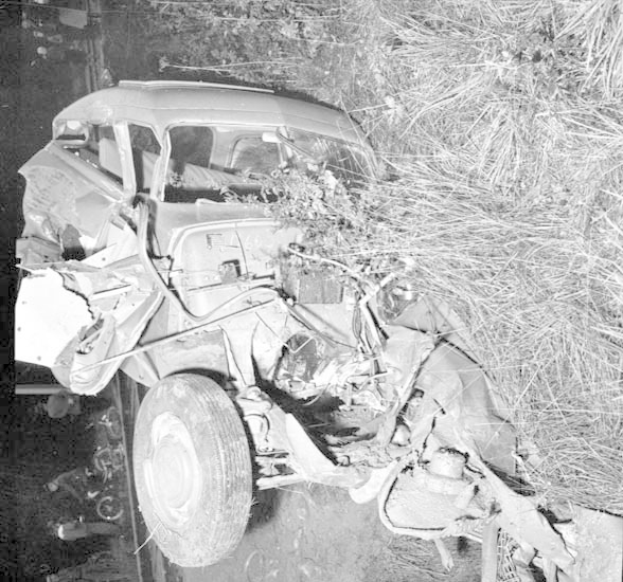 Schwarz-weißes Foto eines stark beschädigten Autos mit eingedellter Front und zersplitterten Fenstern am Straßenrand, mit einem Baum und verstreuten Gegenständen im Hintergrund.