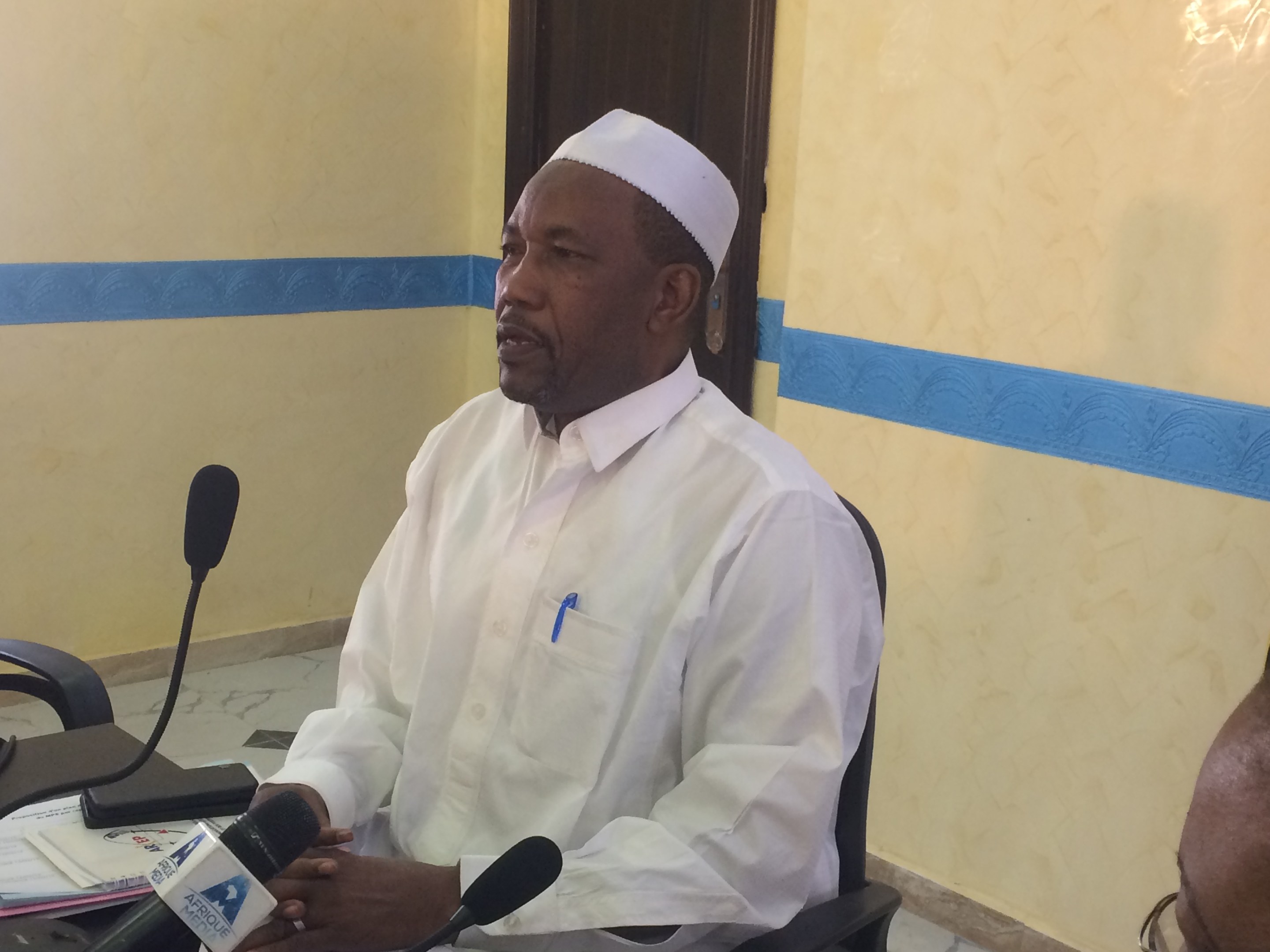 Ein Mann in weißem Anzug und Hut sitzt an einem Tisch mit Mikrofonen, spricht, während Papiere und Gegenstände auf dem Tisch liegen, und ein weiterer Mensch mit Brille rechts von ihm in einem Raum mit Wand und Tür im Hintergrund.