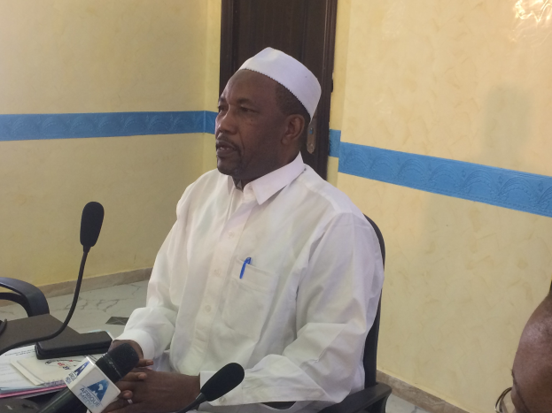 Ein Mann in weißem Anzug und Hut sitzt an einem Tisch mit Mikrofonen, spricht, während Papiere und Gegenstände auf dem Tisch liegen, und ein weiterer Mensch mit Brille rechts von ihm in einem Raum mit Wand und Tür im Hintergrund.