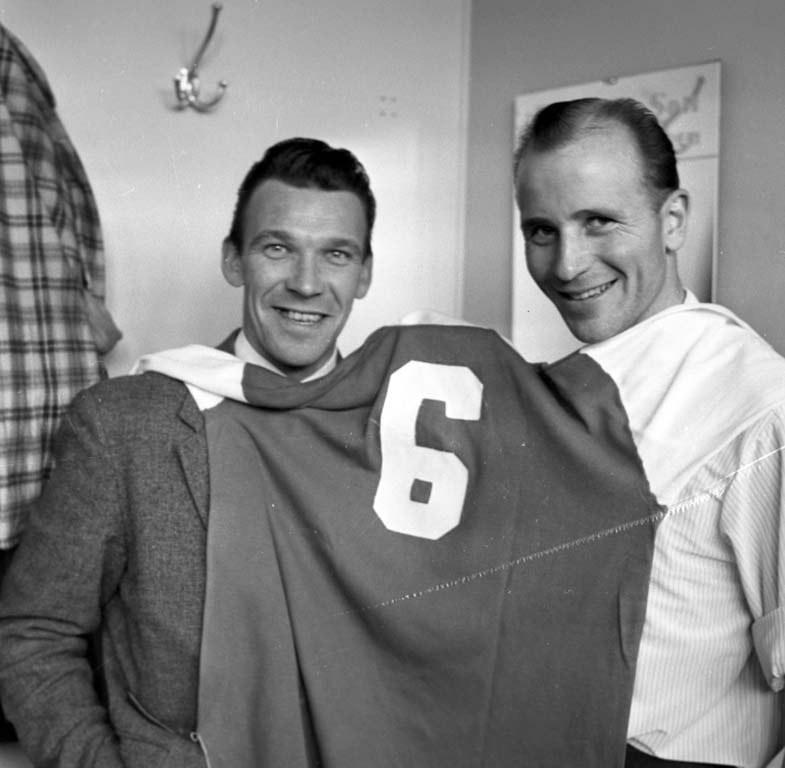 Zwei Männer in Schwarz und Weiß stehen nebeneinander und lächeln, während sie ein Fußballtrikot hochhalten; eine Wandtafel und ein Kleiderbügel sind im Hintergrund zu sehen.