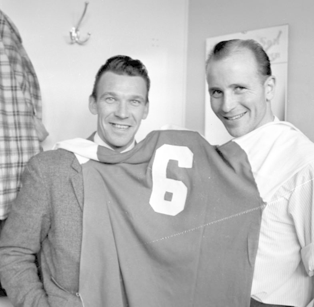 Zwei Männer in Schwarz und Weiß stehen nebeneinander und lächeln, während sie ein Fußballtrikot hochhalten; eine Wandtafel und ein Kleiderbügel sind im Hintergrund zu sehen.