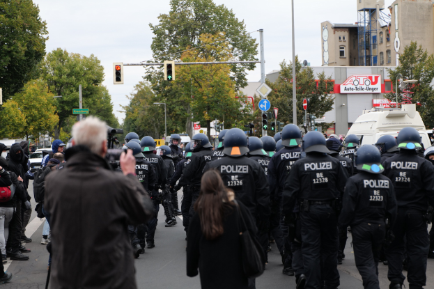 Eine Gruppe von Menschen geht auf einer Straße mit Polizeibeamten, einige tragen Helme und haben Taschen oder halten Kameras, mit Fahrzeugen, Verkehrszeichen, Schildern, Bäumen, Gebäuden und einem klaren blauen Himmel im Hintergrund.