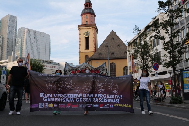 Eine Gruppe von Menschen in Masken, die eine Straße entlanggehen und ein Banner mit Text und Bildern halten, mit einem geparkten Auto auf der linken Seite, Gebäuden, Bäumen, Schildern, Polen, einem Kirchturm und einem klaren blauen Himmel im Hintergrund.