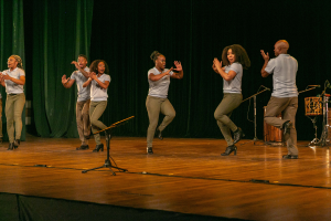 Eine Gruppe von Menschen auf einer Bühne mit Mikrofonen, Musikinstrumenten und einem Vorhang im Hintergrund, die Hände klappend in Feierlaune, die wie ein Tanzteam aussehen.