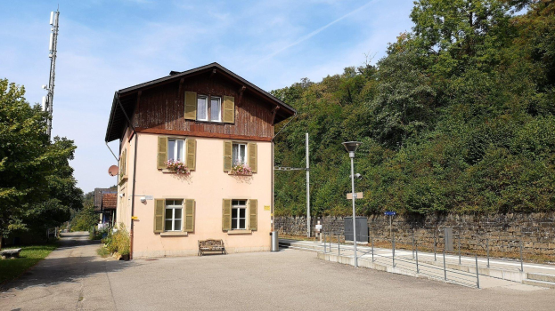 Ferienhaus zum Verkauf in Baden-Württemberg, Deutschland, mit einem Gebäude mit vielen Fenstern, einer vorderen Mauer, Straßenmöbeln, Grünfläche und einem bewölkten Himmel.
