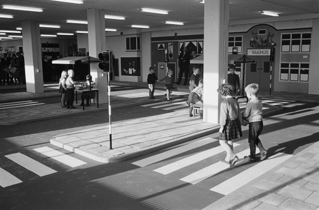 Schwarzes und weißes Foto von Kindern, die eine Straße an einer Ampel überqueren, mit Menschen auf Bänken, einem Verkehrssignalmast im Vordergrund und Säulen mit Deckenlampen im Hintergrund.