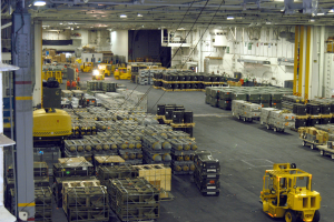 Ein großes Lagerinnere mit zahlreichen Trucks, Containern, Menschen, Lampen, Stangen und anderen Gegenständen, mit einem prominenten Rad.