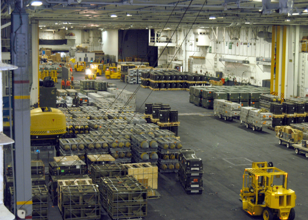 Ein großes Lagerinnere mit zahlreichen Trucks, Containern, Menschen, Lampen, Stangen und anderen Gegenständen, mit einem prominenten Rad.