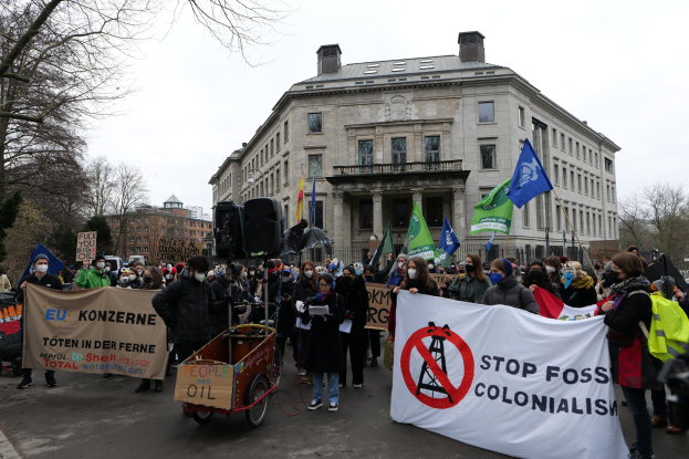 Eine große Gruppe von Menschen marschiert bei einer Demonstration gegen fossile Brennstoffe, trägt Schilder und Fahnen und hat ein Fahrzeug im Vordergrund und Gebäude, Bäume und einen klaren blauen Himmel im Hintergrund.