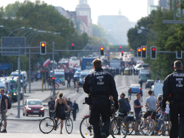 Zwei Polizeibeamte neben einer Gruppe von Radfahrern auf einer Straße mit Fahrzeugen, Verkehrszeichen, Bäumen, Gebäuden und einem klaren blauen Himmel.