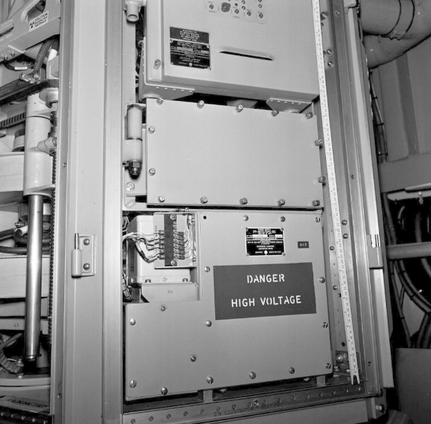 Ein Schwarz-Wei├č-Foto eines elektrischen Schaltkastens mit einer Gefahrenzeichen, umgeben von verschiedenen Maschinen und Rohren.