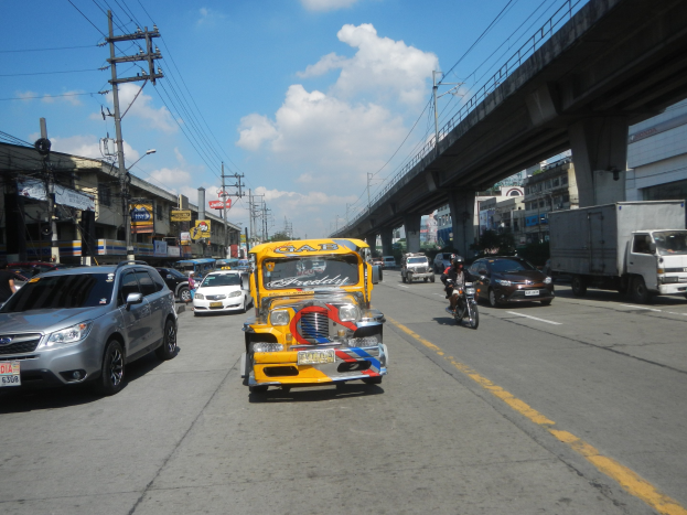Ein gelbes Taxi fährt auf einer Straße mit hohen Gebäuden, Strommasten mit Drähten und einer Brücke im Hintergrund unter einem bewölkten Himmel, mit einem Wasserzeichen.