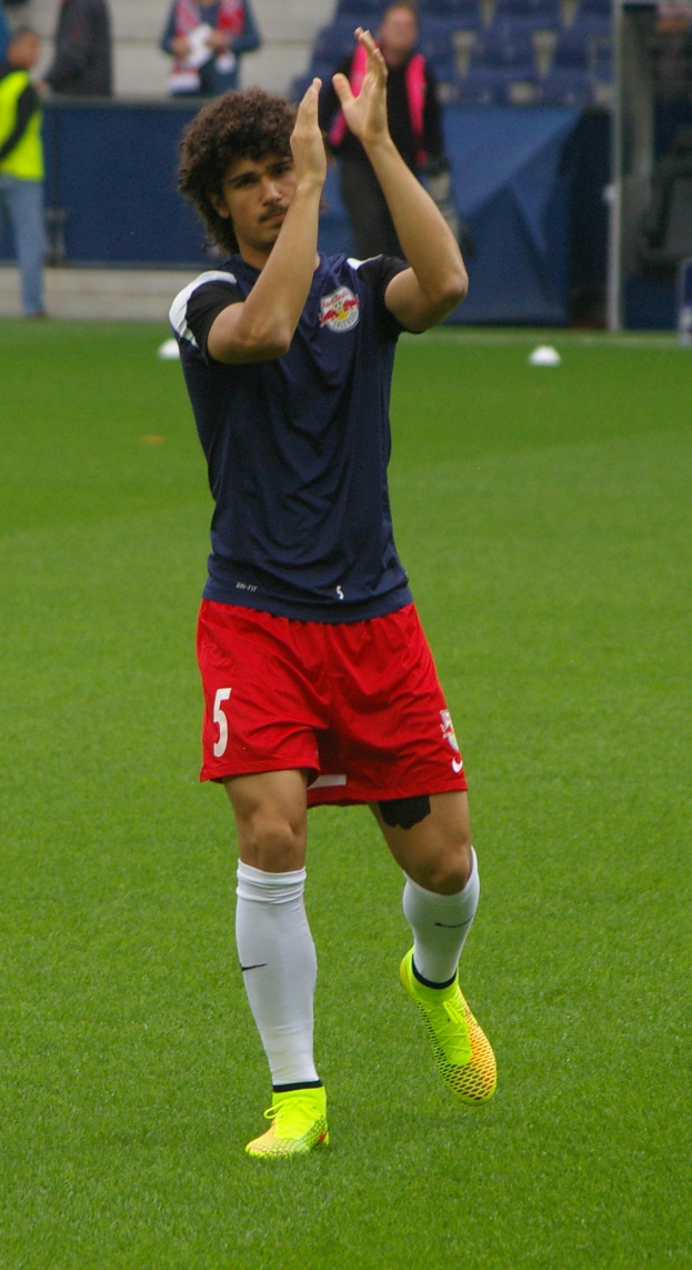 Ein Mann in einem blauen Hemd und roten Shorts steht auf einem Fuballfeld, bereit zum Spielen, mit einer Gruppe von Menschen im Hintergrund.