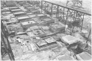 Ein Schwarz-Weiß-Foto einer Baustelle mit einer Brücke im Hintergrund, mit verstreuten Gebäuden, Metallstäben und anderen Gegenständen und Text am unteren Rand.