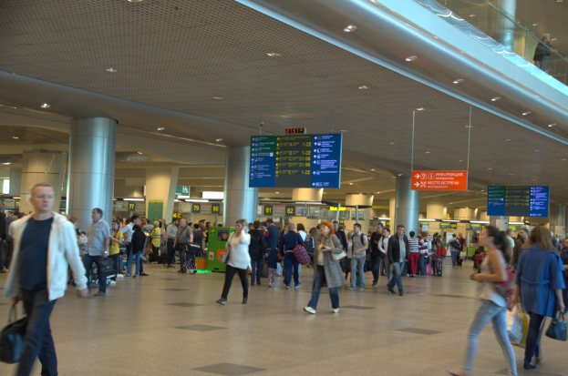 Menschen gehen durch einen Flughafen mit überkopf-Schildern, Säulen, Deckenlampen und einer Glasbrüstung auf der rechten Seite.