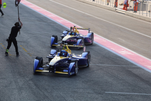 Zwei Formel-E-Autos auf einer Rennstrecke, ein Mann hält eine Fahne daneben, mit ein paar Leuten links und Geländern mit mehr Leuten im Hintergrund.