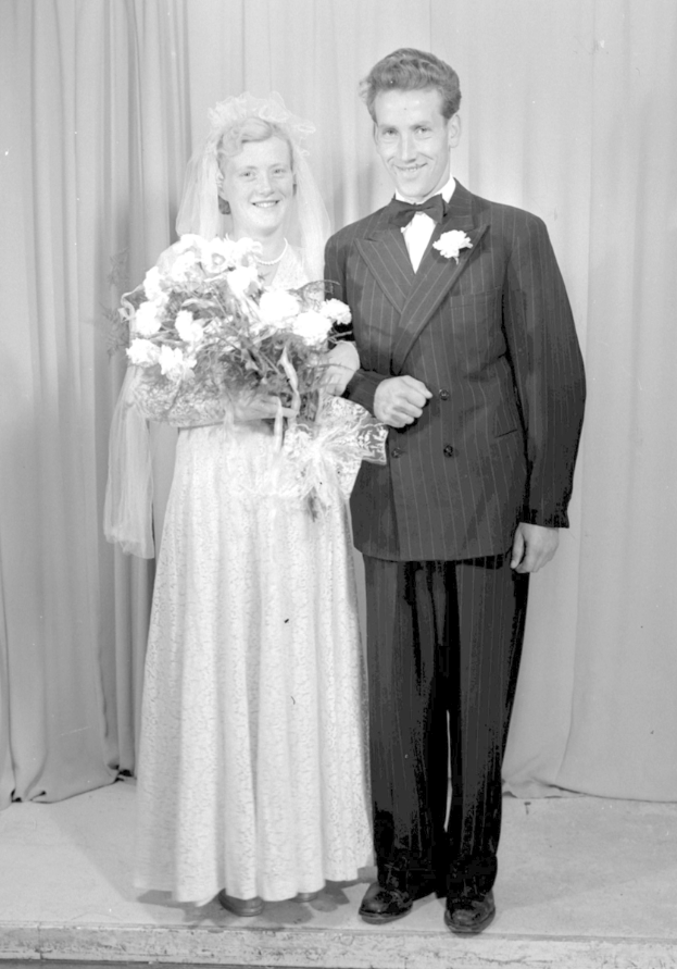 Ein Mann und eine Frau stehen nebeneinander auf einer Bühne, lächeln und die Frau hält einen Blumenstrauß, und ein Vorhang ist hinter ihnen zu sehen.