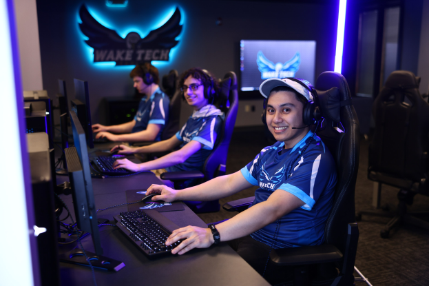 Gruppe von Menschen an einem Schreibtisch mit Computern, Kopfhörern und strahlenden Gesichtern vor Monitoren, Tastaturen und Mäusen, mit einem Logo und Text an der Wand dahinter und Stühlen rechts.