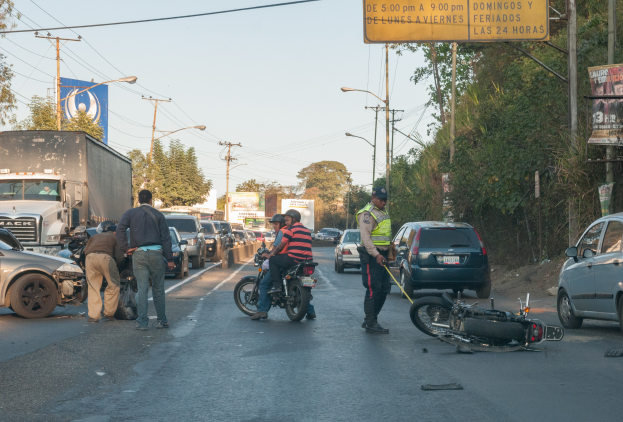 Eine Gruppe von Menschen steht um ein verunglücktes Motorrad auf der Straße herum, umgeben von mehreren Fahrzeugen, darunter ein Lastwagen, und einer Kulisse aus Bäumen, Pfosten, Lampen und Schildern unter dem Himmel.