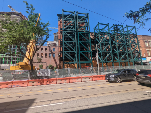 Große Metallbrücke im Bau auf einer Baustelle mit parkenden Autos, einem Zaun, Bäumen, Gebäuden, einem Kran und Himmel im Hintergrund.