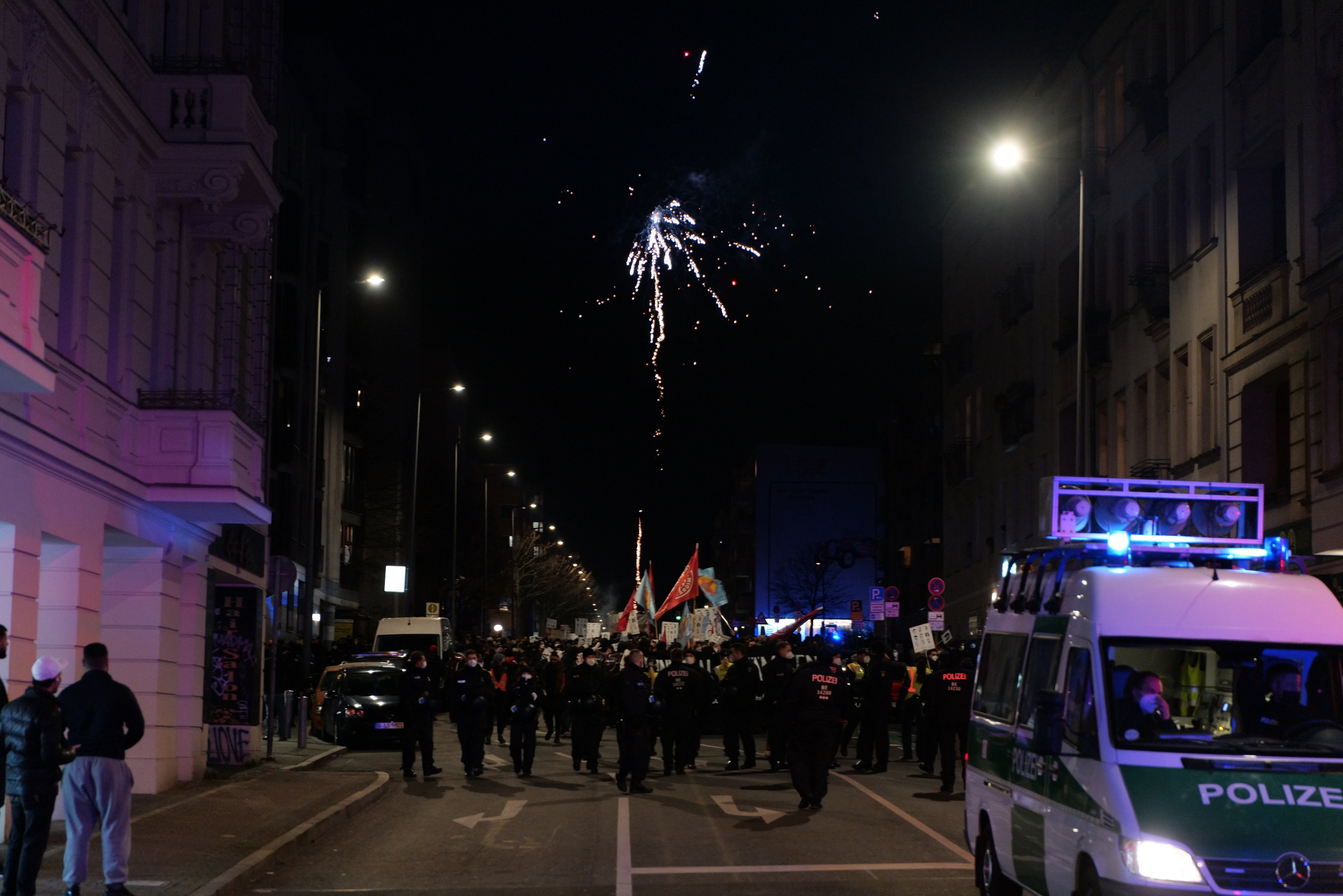 Eine Gruppe von Menschen geht nachts eine Straße entlang, mit Straßenlaternen, Feuerwerk, Fahrzeugen, Gebäuden und Flaggen im Hintergrund.