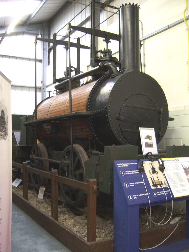 Alte Dampflokomotive in einem Museum mit Informationsschildern, einem Zaun, Steinboden, Banner, Wandrohren und Deckenbeleuchtung ausgestellt.
