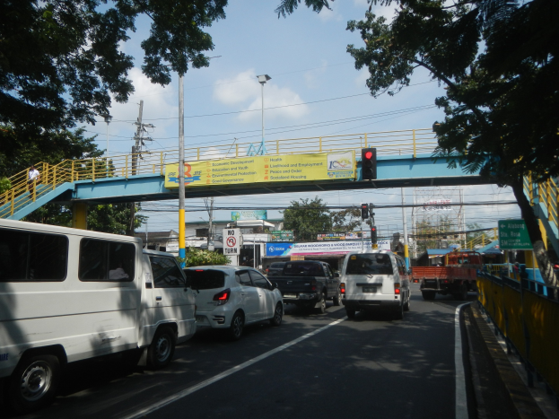 Vollständige Straße mit Fahrzeugen, eine Brücke mit Geländern und Treppen, Laternen, Verkehrsampeln, Texttafeln, Bäume, Gebäude und ein bewölkter Himmel.