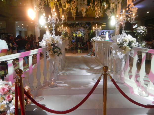 Eine Hochzeitszeremonie in einem Einkaufszentrum mit weißem Teppich, Geländern, dekorativen Seilen, Blumenarrangements, Kronleuchtern und Deckenleuchten, bei der viele Menschen anwesend sind, einige tragen Taschen.