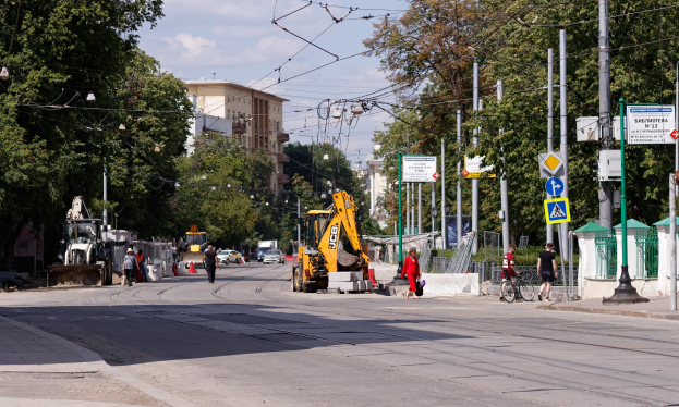 Stadtstraße mit einer Baustelle, Fahrzeugen, Fußgängern, einem Radfahrer, Verkehrszeichen, Strommasten, Bäumen, Gebäuden und einer bewölkten Himmel