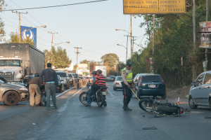 Eine Gruppe von Menschen steht um ein verunglücktes Motorrad auf der Straße mit mehreren Fahrzeugen, darunter ein Lastwagen, und einem Hintergrund aus Bäumen, Pfählen, Lampen und Schildern unter dem Himmel.